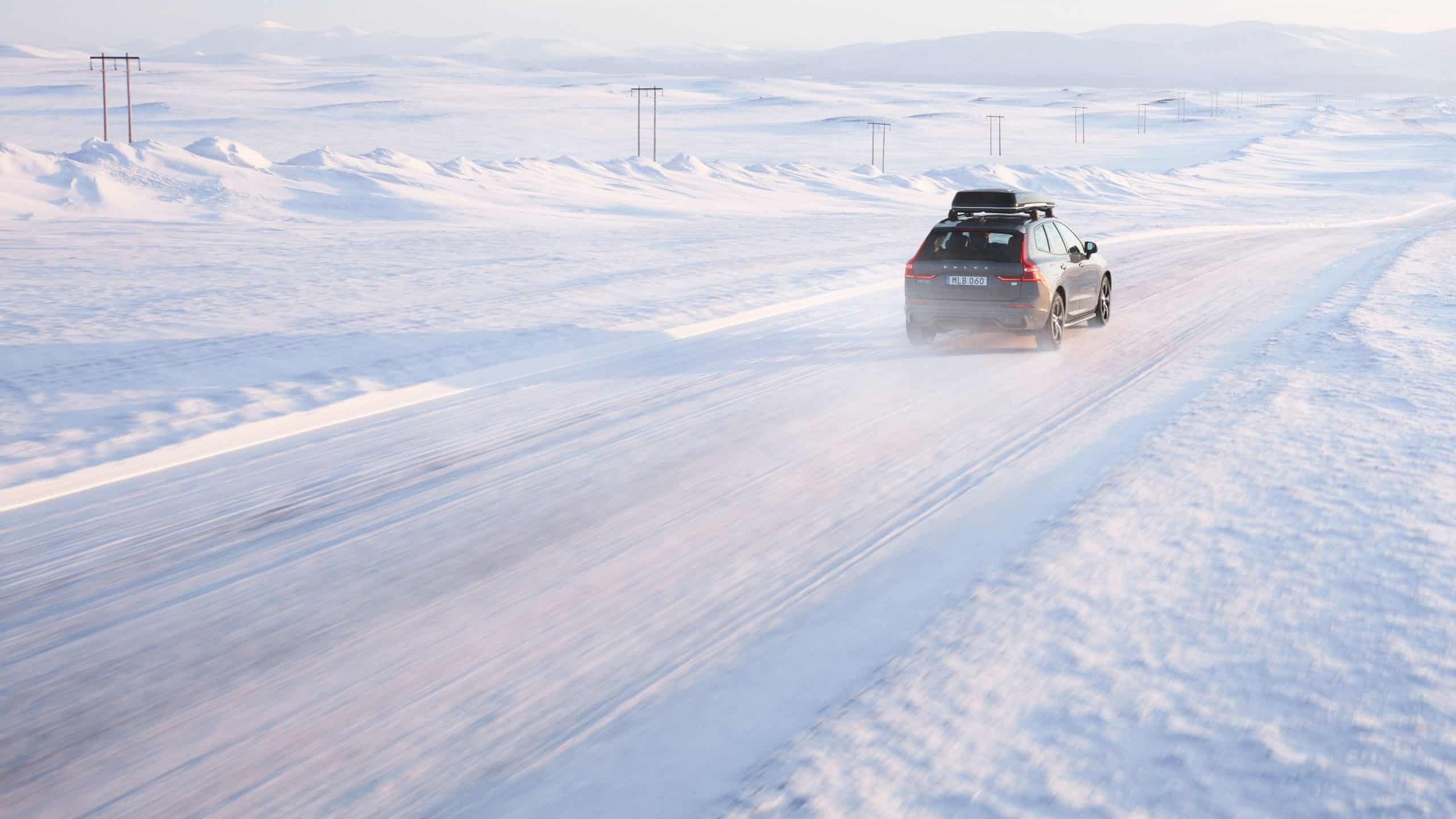 En bil åker i ett vinterlandskap. Det är snö på vägen och snön yr kring vinterdäcken,