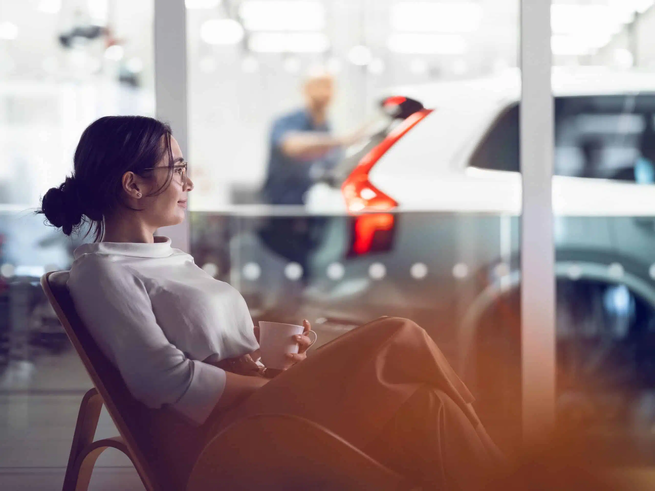 En kvinna sitter med en kopp kaffe i kundytan nära verkstaden. En bil och en servicetekniker syns i bakgrunden.